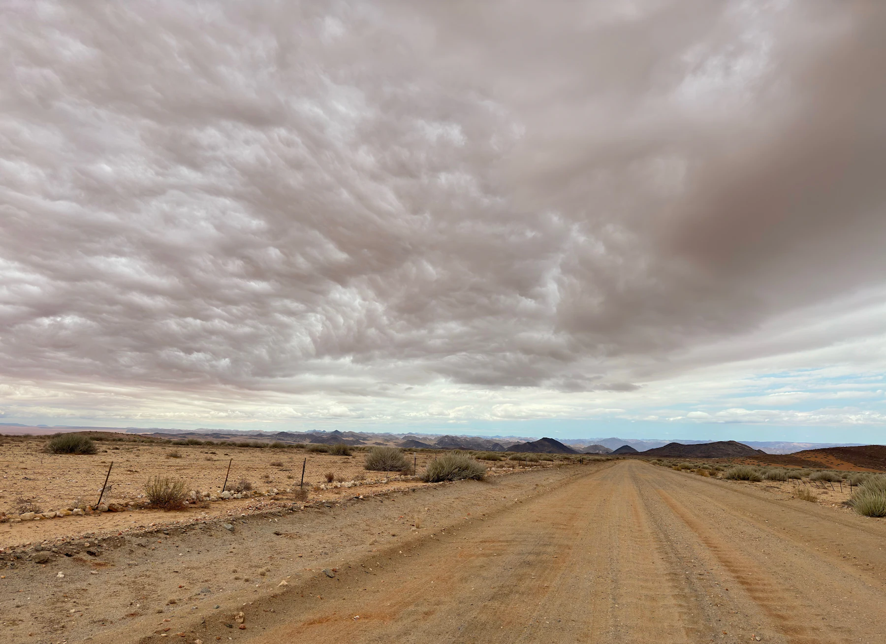 A straight gravel road running across a flat desert towards low hills under a cloudy sky